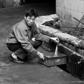 Ouvrier chinois prenant de l'eau dans une usine de fabrication de pièces métalliques. Made in china. Conditions de travail difficiles, conditions précaires. L'ouvrier est accroupi à même le sol. Usine en Chine. 
Noir et blanc.